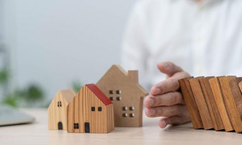 A man’s hand stops wood blocks from falling on a wooden model house